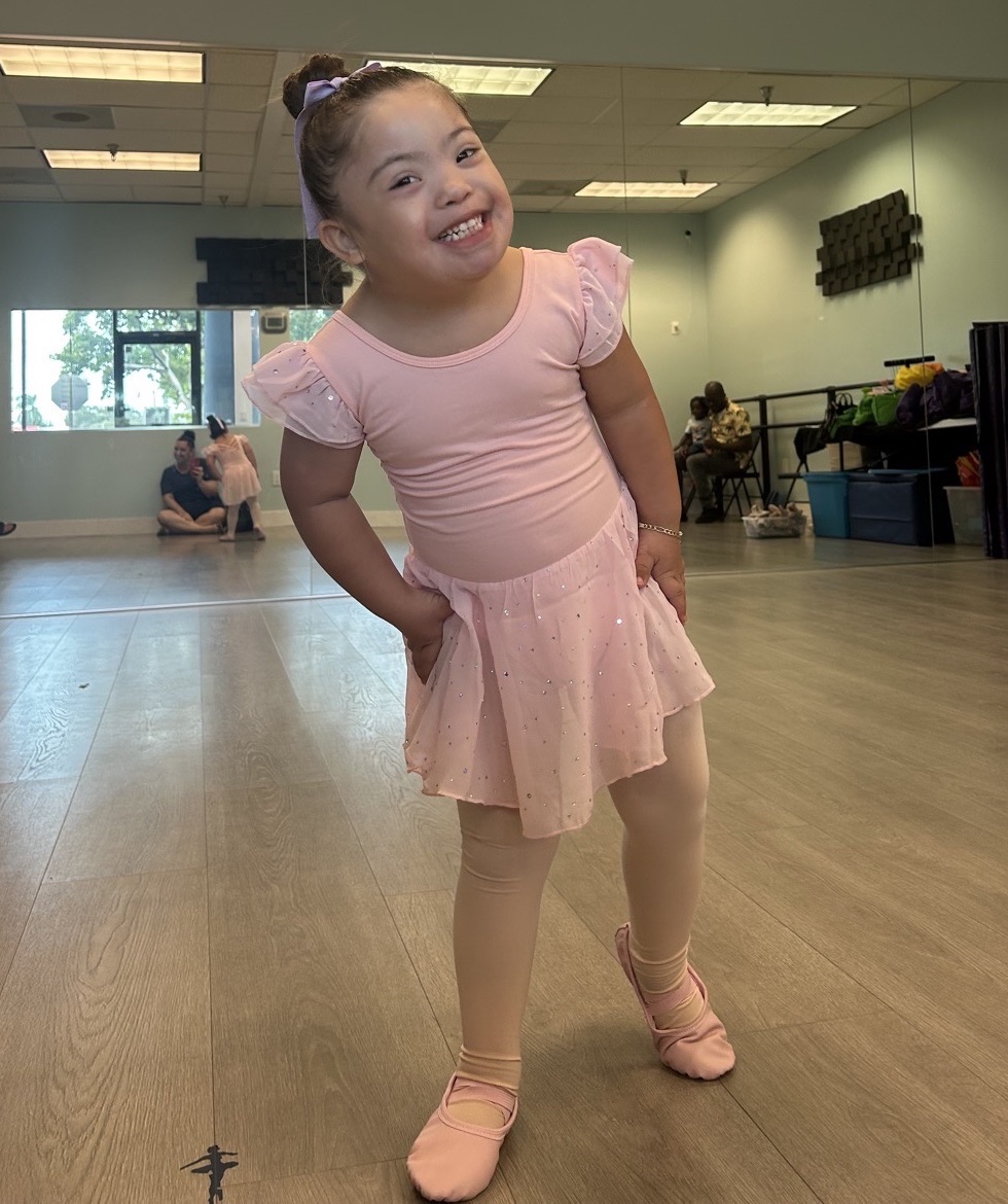 A young ballerina in a pink leotard smiling in a dance studio