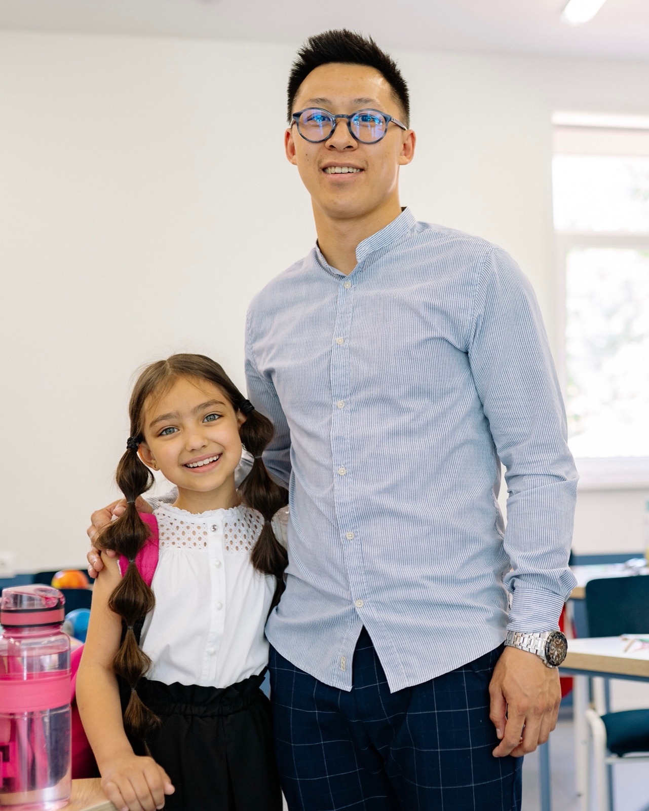 A staff member standing with a young girl in a bright classroom, both smiling